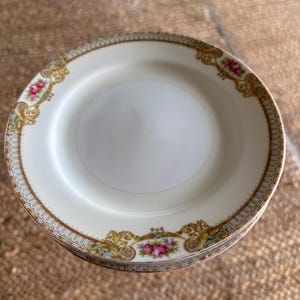 May include: A stack of white porcelain plates with a decorative floral and gold rim. The plates feature pink roses and ornate gold filigree accents. The rim has a delicate blue and gold border. These plates are suitable for serving food.