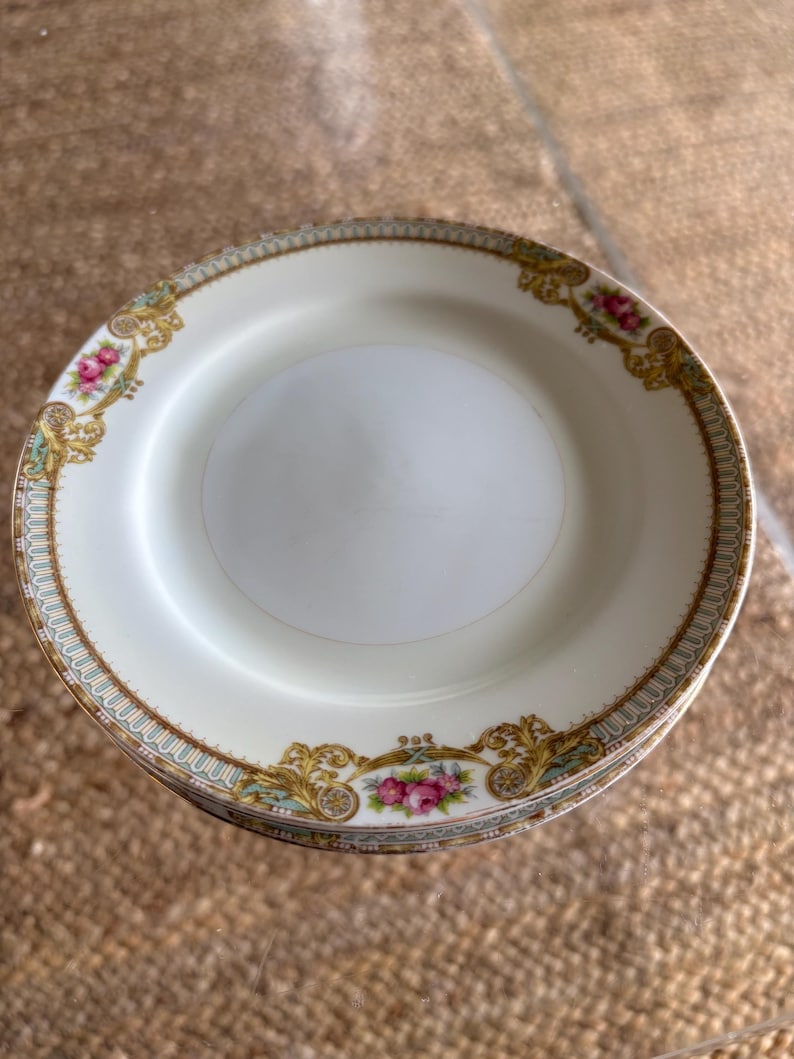 May include: A stack of white porcelain plates with a decorative floral and gold rim. The plates feature pink roses and ornate gold filigree accents. The rim has a delicate blue and gold border. These plates are suitable for serving food.