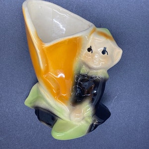 May include: A ceramic planter shaped like a cartoon elf with a green, orange, and black color scheme. The elf is holding a pot with a wide opening.