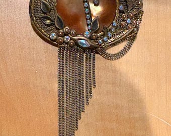 May include: An oval-shaped, antique-style brooch with a copper-colored center and a decorative, textured gold-tone border. The brooch features dangling chains and is embellished with small, sparkling crystals and floral accents.