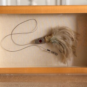 May include: A framed vintage fishing lure. The lure is silver with a red accent and a cluster of tan feathers. It is mounted on a cream-colored fabric within a light brown wooden frame. The lure is attached to a thin silver wire.