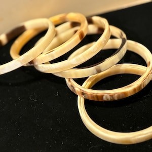 May include: Six ivory-colored bangles with brown marbling are stacked on a black surface. The bracelets have a smooth, polished finish and a circular design. The natural variations in color create a unique look.
