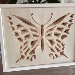 May include: A framed, layered paper art piece featuring a butterfly design. The artwork showcases a layered effect with varying shades of brown and beige, creating depth. The butterfly is centered within a white wooden frame, suitable for wall decor.