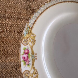 May include: Cream-colored plate with a decorative border featuring gold filigree, teal accents, and pink floral clusters. The plate has a gold rim and a smooth, glossy surface. A classic design for serving food.