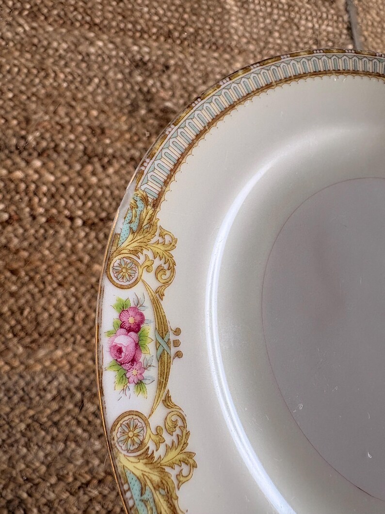 May include: Cream-colored plate with a decorative border featuring gold filigree, teal accents, and pink floral clusters. The plate has a gold rim and a smooth, glossy surface. A classic design for serving food.