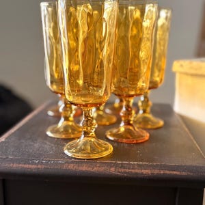 May include: A set of six amber-colored water goblets. Each glass features a textured, rippled design on the bowl and a stemmed base. The glasses are arranged on a dark, distressed wooden surface, showcasing their vintage aesthetic.