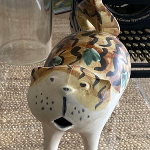 May include: A ceramic cat figurine with brown, white, and green painted details. The cat has a large, open mouth and is standing on four legs.