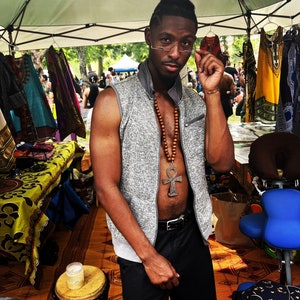 May include: A man wearing a gray vest with a brown wooden beaded necklace and a silver ankh pendant. He is wearing a pair of glasses and has a beard. He is standing in front of a table with colorful fabrics and a blue chair.