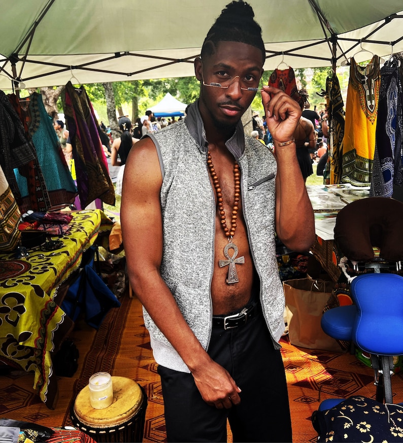 May include: A man wearing a gray vest with a brown wooden beaded necklace and a silver ankh pendant. He is wearing a pair of glasses and has a beard. He is standing in front of a table with colorful fabrics and a blue chair.