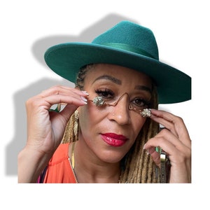 May include: A person wearing a teal green fedora hat and gold-toned glasses with crystal embellishments. The person has long blonde dreadlocks and is wearing a bright orange top.