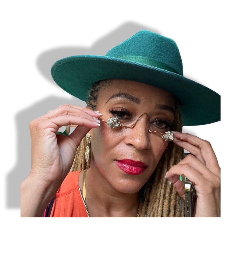 May include: A person wearing a teal green fedora hat and gold-toned glasses with crystal embellishments. The person has long blonde dreadlocks and is wearing a bright orange top.
