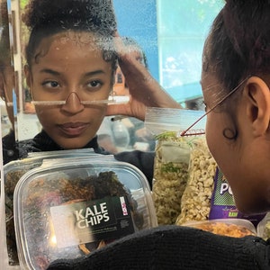 May include: A clear plastic container filled with dark green kale chips. The container has a white label with the text "KALE CHIPS" in black letters.
