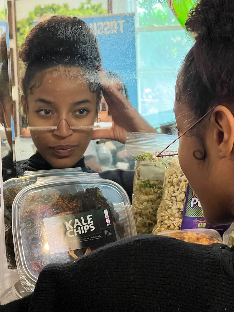 May include: A clear plastic container filled with dark green kale chips. The container has a white label with the text "KALE CHIPS" in black letters.