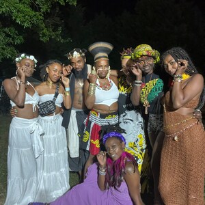 May include: A group of seven people wearing colorful clothing and accessories, including hats, necklaces, and glasses. They are standing in a grassy area at night. The people are smiling and looking at the camera.