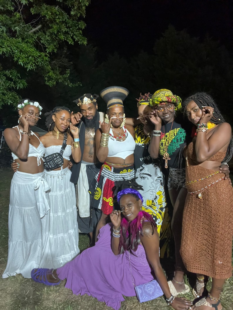 May include: A group of seven people wearing colorful clothing and accessories, including hats, necklaces, and glasses. They are standing in a grassy area at night. The people are smiling and looking at the camera.