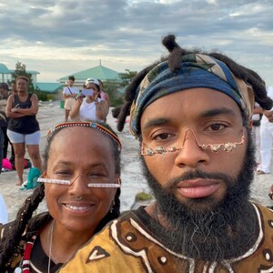 May include: Two people wearing patterned clothing and unique glasses pose for a selfie. The person on the left is wearing a black shirt with a red and white beaded necklace. The person on the right is wearing a brown and white patterned shirt with a blue bandana. Both people are smiling.