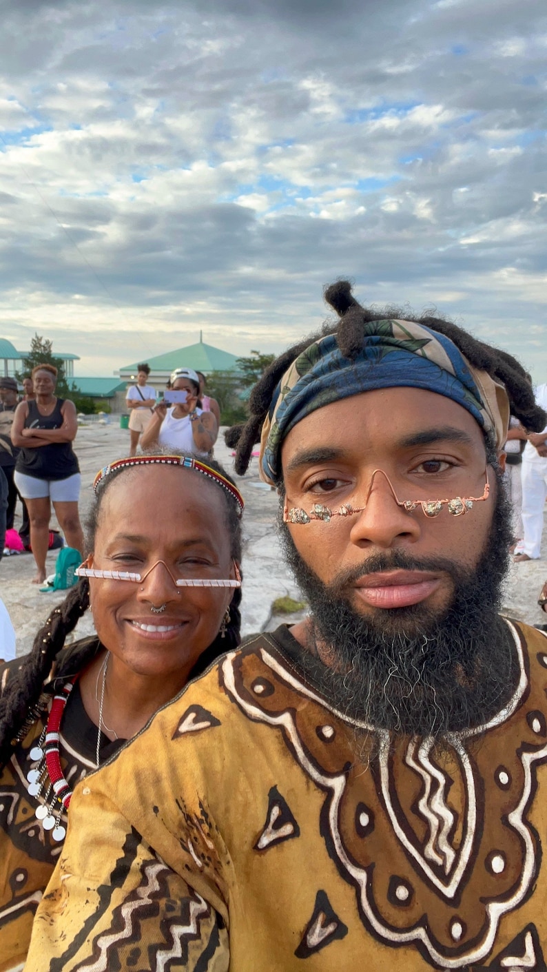 May include: Two people wearing patterned clothing and unique glasses pose for a selfie. The person on the left is wearing a black shirt with a red and white beaded necklace. The person on the right is wearing a brown and white patterned shirt with a blue bandana. Both people are smiling.