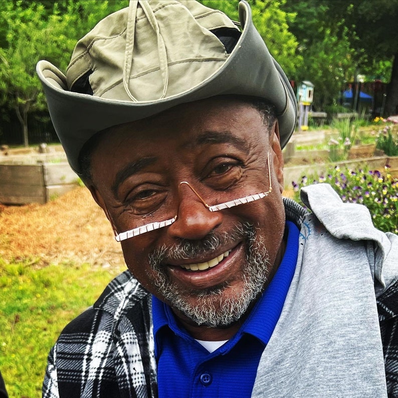 May include: A man wearing a gray and beige cowboy hat with a white brim, a blue collared shirt, and a gray sweater. He is smiling and wearing glasses with a white frame.
