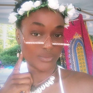 May include: A person wearing a white flower crown, a nose ring, and a necklace with white shell beads. They are also wearing a pair of wire-framed glasses with a pink and white design.