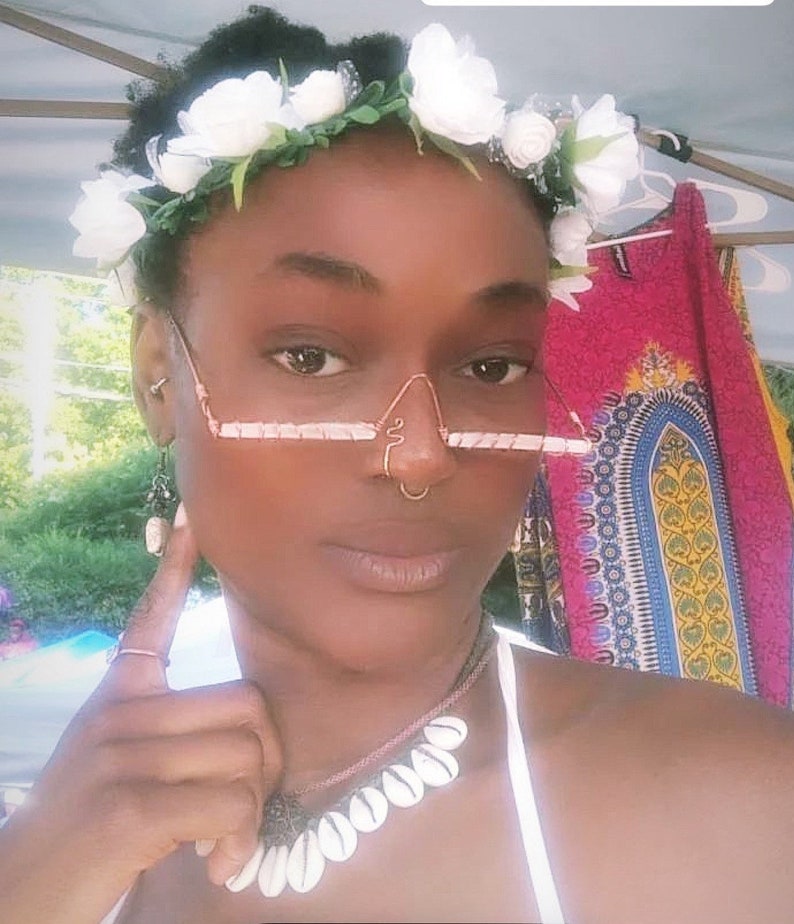 May include: A person wearing a white flower crown, a nose ring, and a necklace with white shell beads. They are also wearing a pair of wire-framed glasses with a pink and white design.