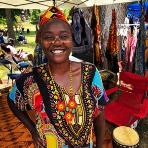 May include: A woman wearing a colorful patterned dress and a beaded necklace smiles at the camera. She is standing in front of a table with various colorful fabrics hanging on it. There is a red folding chair and a wooden drum in the background.