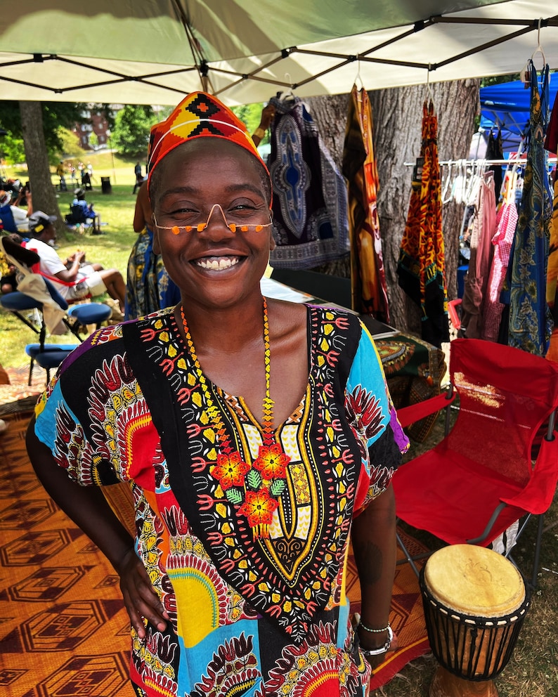 May include: A woman wearing a colorful patterned dress and a beaded necklace smiles at the camera. She is standing in front of a table with various colorful fabrics hanging on it. There is a red folding chair and a wooden drum in the background.
