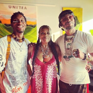 May include: Three people pose for a photo. The person in the middle is wearing a red dress with white floral patterns. The person on the left is wearing a white shirt with a black and white illustration and a yellow strap. The person on the right is wearing a white shirt with a green, black, and gold necklace and a black and green camouflage pants.