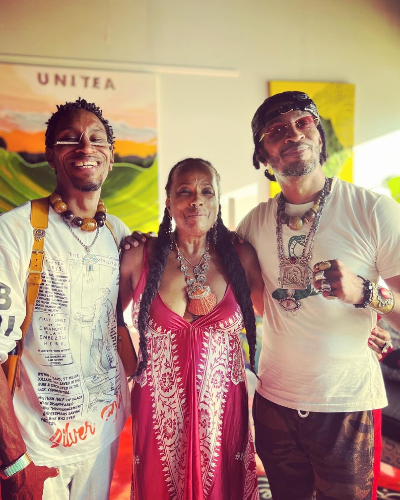 May include: Three people pose for a photo. The person in the middle is wearing a red dress with white floral patterns. The person on the left is wearing a white shirt with a black and white illustration and a yellow strap. The person on the right is wearing a white shirt with a green, black, and gold necklace and a black and green camouflage pants.