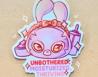 Puede incluir: Una pegatina rosa con un conejito de dibujos animados con una diadema, sosteniendo una botella y un recipiente de crema. La pegatina tiene las palabras "UNBOTHERED. MOISTURIZED. THRIVING" en texto rosa y rojo.