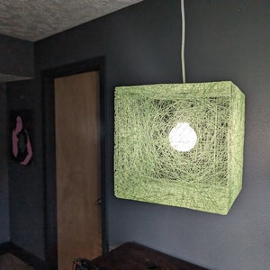 May include: A square-shaped light fixture made of woven green string with a white light bulb inside. The light fixture is hanging from the ceiling.