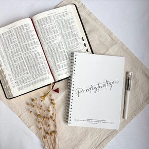 Può includere: Un libro aperto con copertine nere e un segnalibro a nastro rosso accanto a un quaderno a spirale bianco con la parola "Predigtnotizen" in corsivo. Una penna è appoggiata accanto al quaderno. Gli oggetti sono su un tessuto beige.