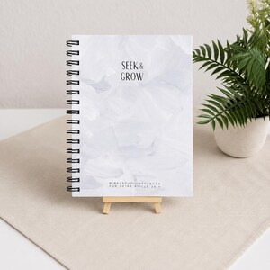 Könnte beinhalten: Ein Spiralnotizbuch mit dem Schriftzug "SEEK & GROW" auf dem Einband. Der Einband hat ein weißes und graues abstraktes Design. Das Notizbuch steht auf einer kleinen Holzstaffelei, mit einer Pflanze in einem weißen Topf im Hintergrund.