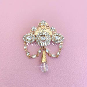 May include: A gold-tone brooch featuring a central oval crystal surrounded by smaller clear rhinestones, with two draped chains of marquise-cut crystals and a dangling teardrop pendant. The text CASABLANCA is visible across the center.