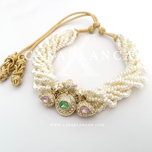 May include: A white pearl choker necklace with a gold and green centerpiece featuring a round green stone and two pink stones. The necklace has a gold tassel on each end.