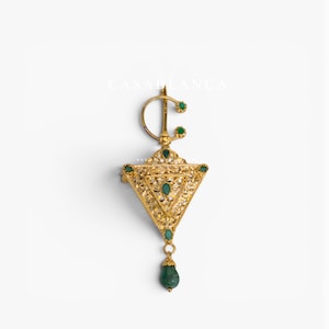 May include: A gold brooch featuring an intricate triangular filigree design adorned with small green stones. The piece includes a circular pin mechanism at the top with two green accents and a dangling teardrop-shaped green stone at the base.