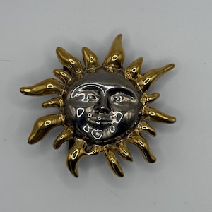 Pode incluir: Broche em forma de sol com rosto prateado e raios dourados. O sol tem um rosto sorridente com detalhes. Os raios dourados se estendem para fora, criando um efeito radiante. O broche é provavelmente feito de metal.