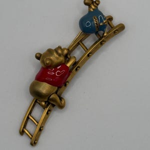 May include: A gold-toned metal brooch featuring a bear character wearing a red shirt climbing a ladder toward a blue pot labeled HUNNY. The bear has a matte gold finish, while the shirt and pot are enameled in vibrant red and blue colors.