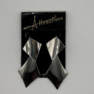 May include: Silver-toned geometric earrings with a two-tiered diamond shape design. The earrings are displayed on a black card with the word "Attractions" in gold script.