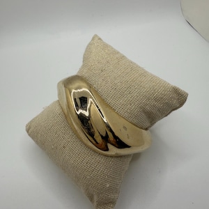 May include: A gold-toned, thick, curved bangle bracelet. The bracelet is smooth and shiny.
