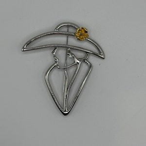 May include: A silver-toned brooch in the shape of a woman wearing a hat with a yellow flower accent.