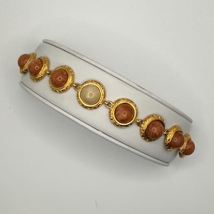 May include: A gold-tone link bracelet featuring a series of circular settings, each containing a smooth, domed cabochon. The bracelet displays six reddish-brown stones and one central, lighter-colored, translucent stone, all framed by ornate gold metal.