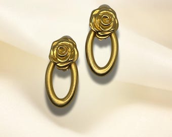May include: A pair of gold-tone earrings featuring a rose design. The earrings are oval-shaped with a rose on top of each oval.