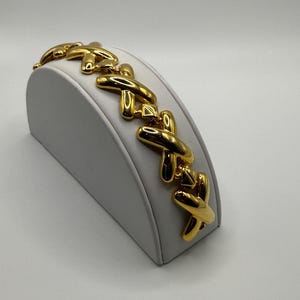 Può includere: Un bracciale a maglie color oro caratterizzato da un motivo ripetitivo di elementi a forma di X lucidi e interconnessi. Il bracciale presenta una superficie liscia e riflettente con un design continuo che avvolge il supporto curvo.