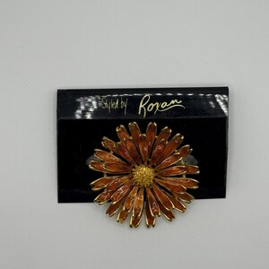 May include: A gold-tone flower brooch with orange enamel petals and a textured gold centre, attached to a black display card featuring the gold text Styled by Roxan. The brooch has multiple layers of slender, pointed petals in a radial pattern.