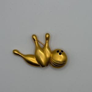 Könnte beinhalten: Eine goldfarbene Metallbrosche in Form einer Bowlingkugel und zwei Bowlingkegeln. Die Bowlingkugel weist drei schwarze kreisförmige Vertiefungen auf, und die zwei Kegel sind aufrecht hinter der Kugel positioniert, mit einem dritten liegenden Kegel.