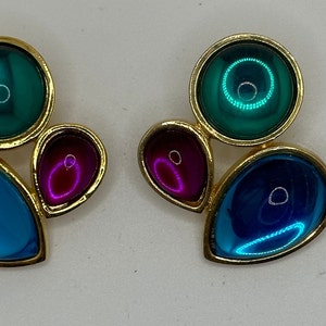 Puede incluir: Un par de pendientes de tono dorado con piedras de vidrio azul, verde y morado. Los pendientes tienen forma de racimo de tres piedras.