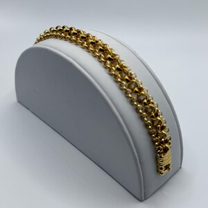 Può includere: Un bracciale color oro caratterizzato da un design a catena a più fili con maglie circolari interconnesse. Il bracciale presenta una finitura lucida e riflettente e include un meccanismo di chiusura rettangolare all'estremità con un motivo strutturato.