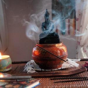May include: A small, black Buddha statue sits atop a red and gold incense burner, with smoke rising from the incense sticks. A lit candle sits on a table next to a book and a wooden incense holder.