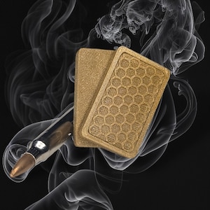 May include: Two gold playing cards with a honeycomb pattern on one side, and a smooth surface on the other side. The cards are partially obscured by a silver bullet and wisps of smoke.