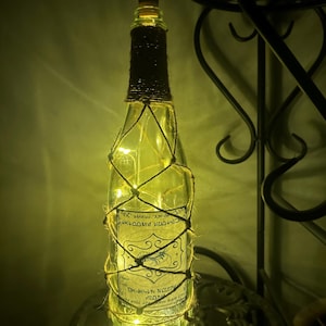 May include: A decorative glass bottle wrapped in twine and illuminated with fairy lights. The bottle has a cork stopper and a label with text and a scorpion illustration. The bottle is on a metal stand.